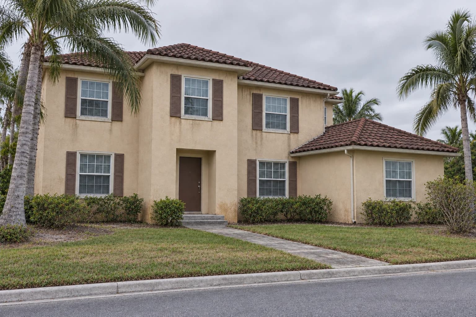 The Palm Residence — Before concept: a Naples house under an overcast sky with faded stucco, a simple recessed entry, weathered roof tiles, a worn concrete path, and patchy lawn
